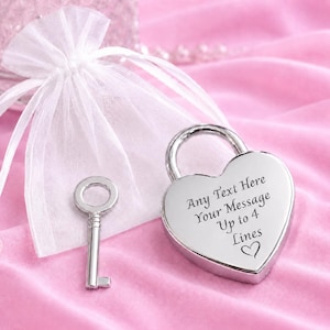 May include: A silver-coloured heart-shaped padlock and a matching key. The padlock features the engraved text: Any Text Here Your Message Up to 4 Lines, followed by a small heart symbol. A white sheer drawstring pouch is positioned behind the items.
