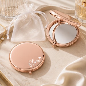 May include: Two round rose gold compact mirrors with a polished metallic finish. One mirror is closed and features the white printed name Olivia with a small heart icon. The other mirror is open, revealing the reflective glass surface. A white organza bag.