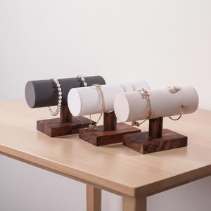 May include: Three jewelry display stands with dark gray, white, and beige cylindrical tops. Each stand has a dark wood base and holds necklaces and bracelets. The stands are on a light wood table.