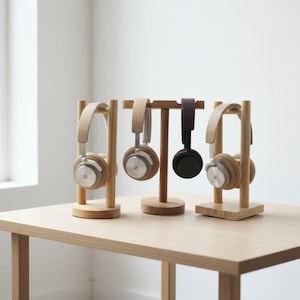 Könnte beinhalten: Kopfhörer werden auf Holzständern präsentiert. Die Ständer sind aus hellem Holz gefertigt und haben ein minimalistisches Design. Die Kopfhörer sind in verschiedenen Farben erhältlich, darunter Beige, Schwarz und Silber. Die Anordnung befindet sich auf einem hellen Holztisch.