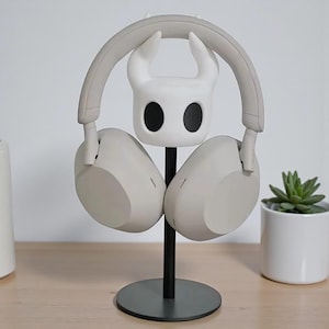 Op de afbeelding: Een wit en grijs hoofdtelefoonstandaard met een gestileerd karakterhoofd, met een paar grijze hoofdtelefoons. De standaard heeft een zwarte paal en een cirkelvormige basis. Een kleine vetplant in een pot is zichtbaar op de achtergrond.