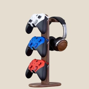 Pode incluir: Um suporte de madeira para acessórios de jogos com três comandos de jogos brancos, azuis e vermelhos, e um par de auscultadores pretos e prateados. O suporte é feito de madeira escura e tem uma base arredondada.