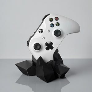 Op de afbeelding: Een witte Xbox-controller op een zwarte geometrische standaard. De controller heeft gekleurde knoppen en zwarte joysticks. De standaard heeft een facetontwerp, wat een moderne esthetiek creëert. De controller is omhoog gekanteld.