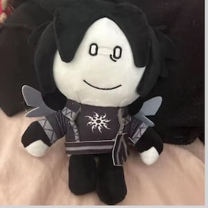 Puede incluir: Una muñeca de peluche con una cara blanca, cabello negro y un atuendo negro y gris. La muñeca tiene una cara sonriente con dos ojos pequeños y un diseño similar al sol en el pecho. Tiene pequeñas alas grises.