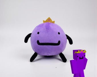 May include: A round purple plush toy with a soft texture, featuring two small black arms and two black legs. The plush has a simple black embroidered mouth and a small yellow crown on top. Next to it is a small pixelated purple character graphic.