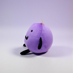 May include: A round, purple plush toy with a soft, fuzzy texture. It features a simple black embroidered face with two small circular eyes and a curved mouth. The toy has two black, elongated limbs extending from the sides and a small yellow tag.