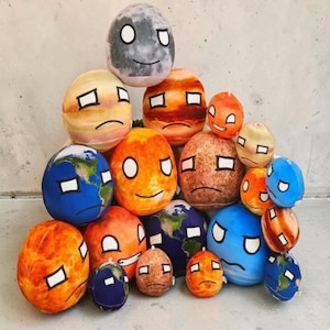 Op de afbeelding: Een verzameling pluche planeetknuffels, elk met een uniek ontwerp dat verschillende hemellichamen vertegenwoordigt. De knuffels hebben verschillende kleuren, waaronder blauw, oranje en grijs, met expressieve gezichten en rechthoekige ogen. De knuffels zijn in een piramidevorm gestapeld.