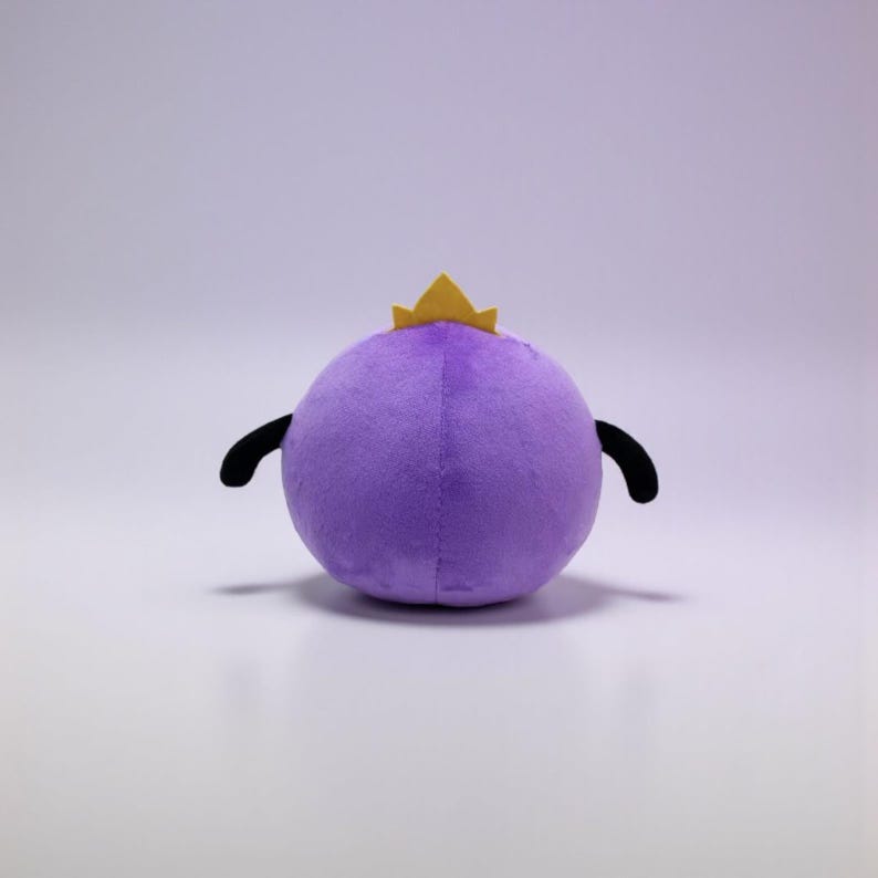 May include: A round purple plush toy featuring a small yellow crown on top and two black, curved appendages extending from the sides. The plush has a soft, textured surface and a spherical shape, presented against a plain, light-colored background.