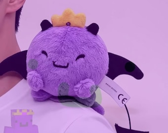 May include: A round purple plush toy featuring a yellow crown, black bat-like wings, and a pointed tail. The creature has a smiling expression with closed eyes, small black horns, and a white tag with the CE mark attached to its side.