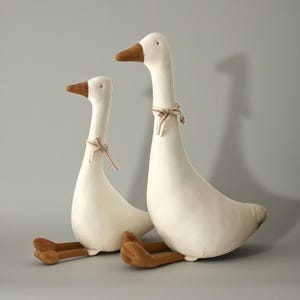 Könnte beinhalten: Zwei Plüschgänse in Weiß mit braunen Schnäbeln und Füßen sitzen. Jede Gans hat ein kleines gestreiftes Band um den Hals gebunden. Die Gänse sind vor einem einfarbigen grauen Hintergrund positioniert.