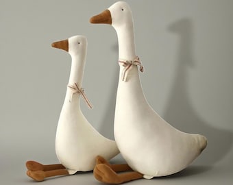 May include: Two plush white geese with brown beaks and feet are seated. Each goose has a small striped ribbon tied around its neck. The geese are positioned against a solid gray background.