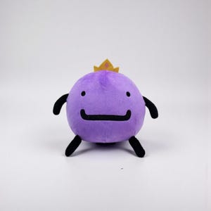 May include: A round purple plush toy featuring a small yellow crown on top, two black eyes, a wide black mouth, and four black limbs. The soft character has a smooth texture and a simple, symmetrical design with a flat base for stability.