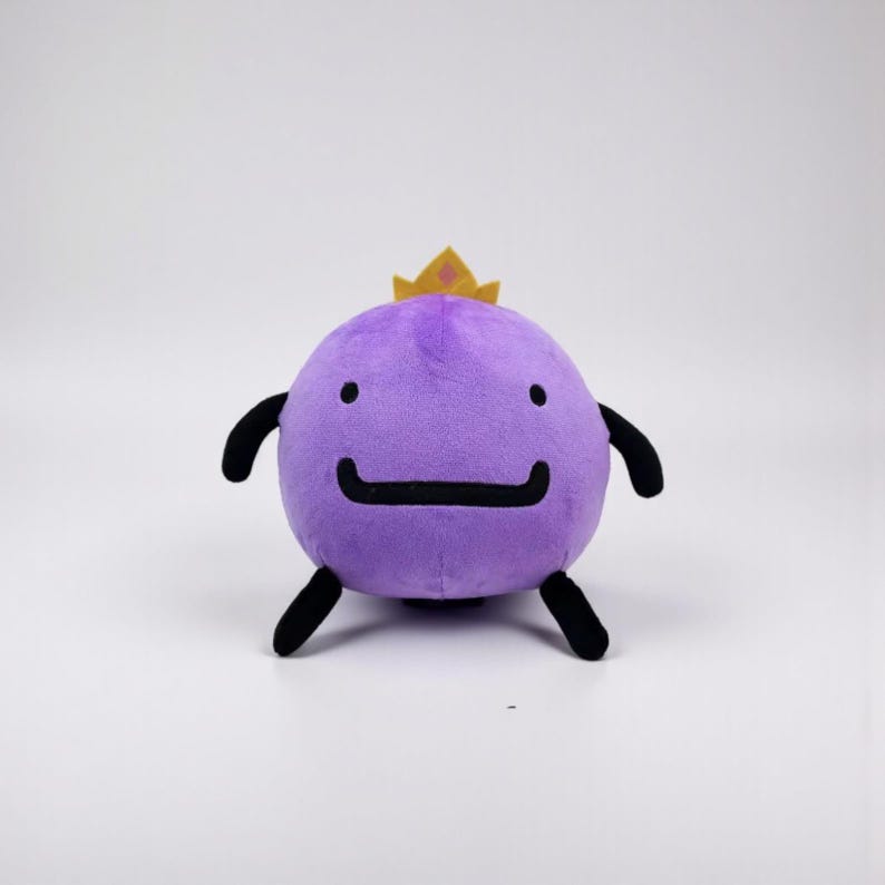 May include: A round purple plush toy featuring a small yellow crown on top, two black eyes, a wide black mouth, and four black limbs. The soft character has a smooth texture and a simple, symmetrical design with a flat base for stability.