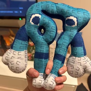 Op de afbeelding: Een blauw pluche speelgoed met een pixelpatroon en witte details. Het speelgoed heeft een abstracte vorm met afgeronde kenmerken. Het ontwerp bevat donkerblauwe en lichtblauwe secties. Het speelgoed wordt in een hand gehouden. Het is een zacht, knuffelbaar item.