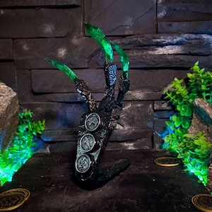 Puede incluir: Una escultura de mano negra estilizada con tres garras verdes puntiagudas. La mano está adornada con tres adornos circulares en forma de moneda. La escultura se exhibe sobre una plataforma negra, con dos monedas doradas cerca.