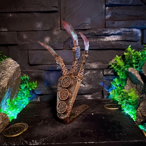 Może przedstawiać: Brązowa, stylizowana rzeźba dłoni z długimi, ostrymi pazurami z czerwonymi końcówkami. Dłoń jest misternie zdobiona okrągłymi wzorami i spoczywa na czarnej, teksturowanej powierzchni. Ozdobne monety umieszczono wokół podstawy, z ciemnym kamiennym tłem.