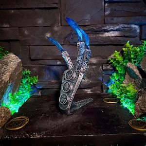 Pode incluir: Uma escultura de fantasia de uma mão com unhas longas e azuis. A mão é prateada e preta com detalhes intrincados. A escultura está sobre uma plataforma preta com moedas decorativas e folhagem verde.