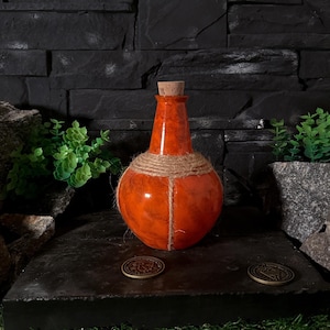 Puede incluir: Una botella de poción de vidrio naranja con tapón de corcho y detalles de cuerda. La botella se encuentra sobre una superficie oscura con dos monedas de bronce. El fondo presenta una pared de piedra negra y vegetación.