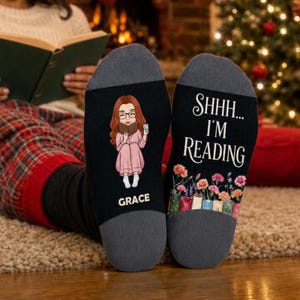 May include: A pair of black socks with gray heels and toes. One sock features a cartoon illustration of a person with long red hair reading a book, with the name GRACE printed below. The other sock displays the text SHHH... I'M READING above a row of colorful books and flowers.