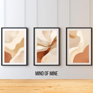 Może przedstawiać: Trzy oprawione abstrakcyjne wydruki artystyczne w neutralnych odcieniach beżu, brązu i rdzy. Każdy wydruk przedstawia unikalny wzór płynących kształtów. Ramki są czarne, a pod nimi znajduje się napis "MIND OF MINE".