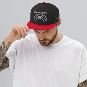 May include: Black and red baseball cap with the text "TI SPACC0!" and two white fists. The cap has a flat brim and a small Italian flag on the side.