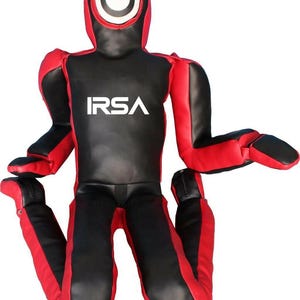 以下が含まれることがあります： 白字で「IRSA」と書かれた赤と黒のトレーニングダミー。 ダミーは、頭、腕、脚を備えた人間の姿に似せて設計されています。 頭には大きな白い円があります。 腕と脚は曲がっています。