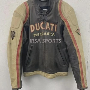Peut inclure: Veste de moto Ducati vintage en cuir gris et beige. La veste présente des rayures rouges sur les manches et le logo Ducati sur la poitrine. Le texte "Meccanica" et "IRSA SPORTS" est également visible.