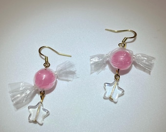 May include: A pair of whimsical earrings featuring pink candy-shaped beads wrapped in clear cellophane. Each earring has a gold-colored hook and a dangling clear star charm. The earrings are designed to resemble wrapped candies.