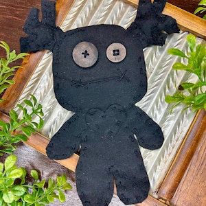 May include: A black felt doll with button eyes and stitched details. The doll has a heart-shaped design on its chest and antler-like shapes on its head. It is displayed on a decorative wooden frame with a glass insert, surrounded by greenery.