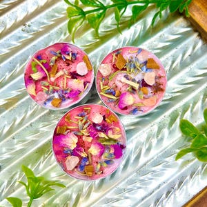 May include: Three round, pink candles with a variety of dried flowers, herbs, and crystals. The candles are in silver containers and sit on a reflective, textured surface. Green foliage is in the background.