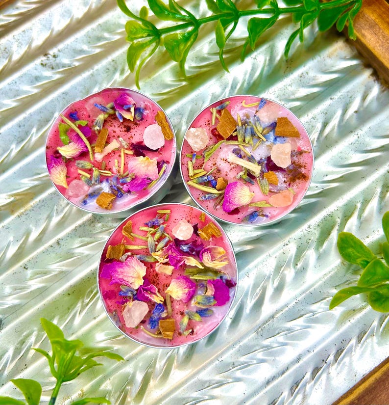 May include: Three round, pink candles with a variety of dried flowers, herbs, and crystals. The candles are in silver containers and sit on a reflective, textured surface. Green foliage is in the background.