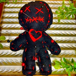 May include: A black voodoo doll with red stitching and a red heart. The doll has pins with red and black heads inserted into it. The doll is on a wooden tray with green foliage.