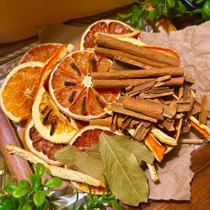 Könnte beinhalten: Ein Holztablett mit getrockneten Orangenscheiben, Zimtstangen, Lorbeerblättern und Nelken. Die Orangenscheiben sind leuchtend orange, die Zimtstangen sind braun. Die Lorbeerblätter sind grün und die Nelken sind klein und dunkel.