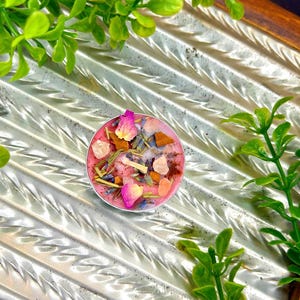 May include: A small, round, pink candle with dried flowers, crystals, and herbs. The candle is in a silver container and is surrounded by green foliage. The candle's surface is decorated with pink rose petals, lavender, and small crystals.