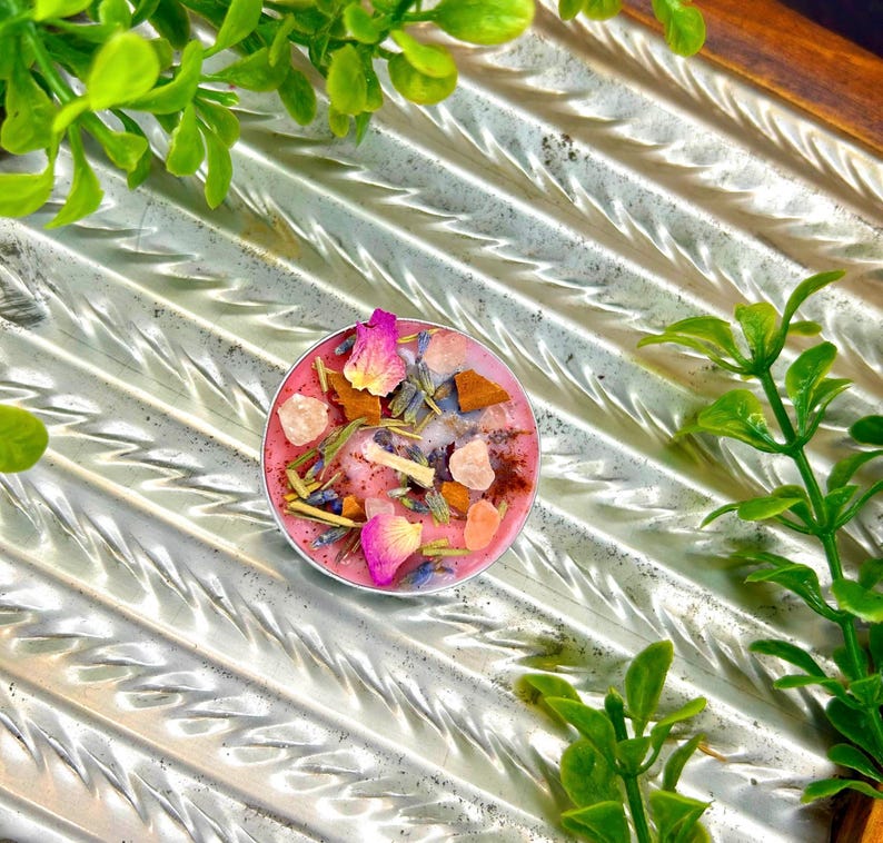 May include: A small, round, pink candle with dried flowers, crystals, and herbs. The candle is in a silver container and is surrounded by green foliage. The candle's surface is decorated with pink rose petals, lavender, and small crystals.
