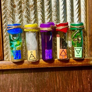 May include: Five glass vials, each representing an element. The vials are sealed with coloured tops and twine. The elements are water, air, purple, fire, and earth, each with a symbol and contents inside the vials.