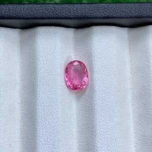 May include: A faceted, oval-shaped pink gemstone, set against a white background within a black jewelry box. The gemstone is a vibrant shade of pink, reflecting light. This image showcases a precious stone, likely for jewelry.