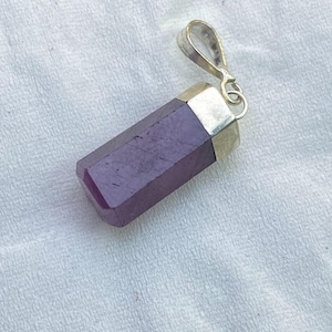 May include: A rectangular purple gemstone pendant with a smooth, polished finish. The stone is set in a silver-toned metal cap that features a small, circular bail at the top for attachment to a necklace chain or cord.