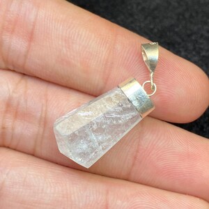 Puede incluir: Un colgante de cristal transparente, facetado y de corte bruto, engastado en una tapa de metal plateado con una anilla a juego. El cristal tiene una forma geométrica irregular y translúcida con fracturas internas visibles y una superficie plana.