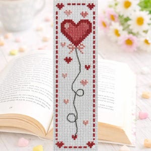 Puede incluir: Marcapáginas de punto de cruz hecho a mano con un diseño de globo de corazón rojo. El marcapáginas tiene un fondo blanco con detalles de corazón rojo y rosa, y una cuerda negra. Los bordes están bordeados con punto de cruz rojo.