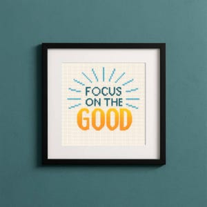 May include: A framed cross-stitch artwork with the words "FOCUS ON THE GOOD" in teal and orange. The artwork is in a white mat and black frame, hanging on a teal wall. The design features a sunburst pattern above the words.