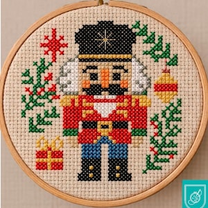 Puede incluir: Bordado de punto de cruz de un soldado Cascanueces en un aro de madera. El soldado lleva un sombrero negro, una chaqueta roja y pantalones azules. Acentos navideños verdes y rojos rodean al soldado, incluyendo un regalo, un copo de nieve y una bola.