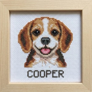 Op de afbeelding: Kruissteekkunstwerk van een glimlachende beagle-hond met bruine, witte en zwarte vacht, omlijst in een licht houten frame. De naam van de hond, "COOPER", is eronder geborduurd.
