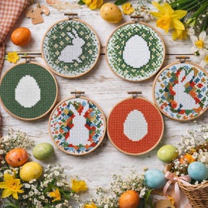 Puede incluir: Seis aros de punto de cruz redondos con diseños de Pascua. Los aros presentan imágenes de huevos y conejos en varios colores, sobre un fondo de madera blanca. Huevos de Pascua y flores rodean los aros.