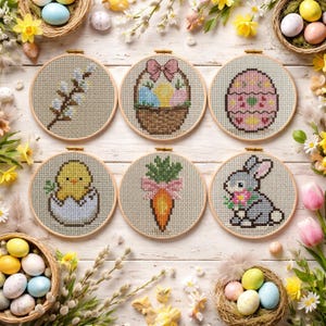 May include: Six round cross-stitch embroidery hoops with Easter-themed designs. The designs include a pussy willow branch, a basket of eggs, a decorated egg, a baby chick, a carrot, and a bunny holding flowers. The hoops are arranged on a white wooden surface.
