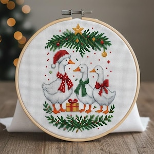 Può includere: Un telaio da ricamo a punto croce con tre oche bianche adornate con accessori festivi. Un'oca indossa un cappello da Babbo Natale e una sciarpa rossa, un'altra una sciarpa verde e la terza un fiocco rosso. Un albero di Natale e una stella in cima.