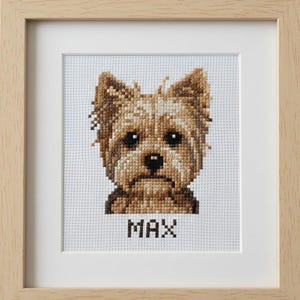Può includere: Ritratto a punto croce di un cane Yorkshire Terrier di nome Max. L'opera d'arte utilizza sfumature di marrone e beige, su uno sfondo chiaro. Il nome del cane, "MAX", è cucito a lettere maiuscole sotto l'immagine. Il pezzo incorniciato è in una cornice di legno chiaro.