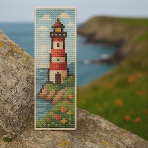 Könnte beinhalten: Ein Kreuzstich-Lesezeichen mit einem Leuchtturm auf einer Klippe. Der Leuchtturm hat einen rot-weiß gestreiften Turm, eine schwarze Spitze und ein gelbes Licht. Die Szene umfasst blaues Wasser, einen blauen Himmel mit Wolken und grünes Gras mit Blumen.
