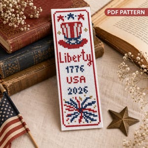 May include: A white cross-stitch bookmark with a red border features patriotic imagery. The design includes an Uncle Sam hat, the word "Liberty," the year "1776," "USA," and the year "2026." A fireworks design is at the bottom.
