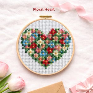 May include: A cross-stitch embroidery hoop featuring a heart design filled with colorful flowers. The heart is composed of red, blue, pink, and yellow cross-stitches on a white background. The text "Floral Heart" is above the hoop.